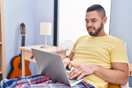 Foto de Hombre latino joven usando portátil sentado en la cama en el dormitorio - Imagen libre de derechos