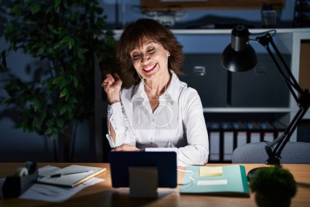 Téléchargez les photos : Femme d'âge moyen travaillant au bureau la nuit avec un grand sourire sur le visage, pointant du doigt vers le côté en regardant la caméra. - en image libre de droit