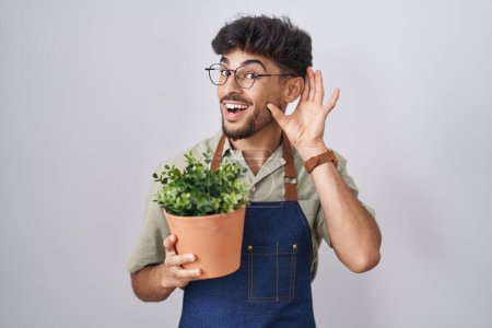 Téléchargez les photos : Homme arabe avec barbe tenant pot de plante verte souriant avec la main sur l'oreille à l'écoute d'une audience à la rumeur ou des commérages. concept de surdité. - en image libre de droit
