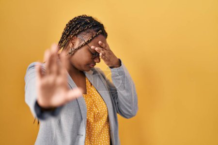Photo for African american woman with braids standing over yellow background covering eyes with hands and doing stop gesture with sad and fear expression. embarrassed and negative concept. - Royalty Free Image