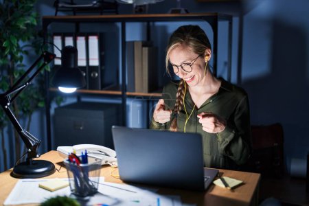Photo for Young blonde woman working at the office at night pointing fingers to camera with happy and funny face. good energy and vibes. - Royalty Free Image