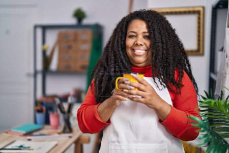 Foto de Mujer afroamericana artista sonriendo confiado bebiendo café en el estudio de arte - Imagen libre de derechos