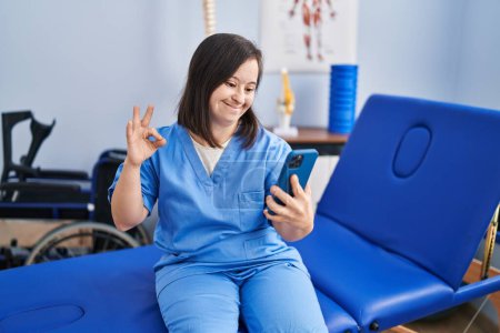 Photo for Hispanic girl with down syndrome working at physiotherapy using smartphone doing ok sign with fingers, smiling friendly gesturing excellent symbol - Royalty Free Image