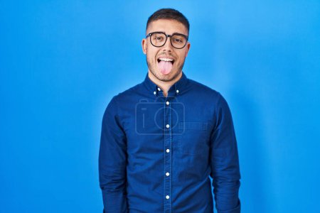 Téléchargez les photos : Jeune homme hispanique portant des lunettes sur fond bleu langue collante heureux avec une expression drôle. concept d'émotion. - en image libre de droit