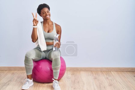 Téléchargez les photos : Femme afro-américaine portant des vêtements de sport assis sur une balle de pilates souriant en regardant la caméra montrant des doigts faisant signe de victoire. numéro deux. - en image libre de droit