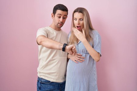 Téléchargez les photos : Jeune couple s'attendant à un bébé debout sur fond rose regardant le temps de la montre inquiet, peur de se faire tard - en image libre de droit