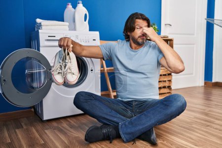 Foto de Hombre de mediana edad sosteniendo zapatillas sucias sentadas en el suelo en la sala de lavandería - Imagen libre de derechos