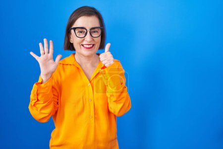 Téléchargez les photos : Moyen Age femme hispanique portant des lunettes debout sur fond bleu montrant et pointant vers le haut avec les doigts numéro six tout en souriant confiant et heureux. - en image libre de droit