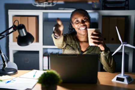 Foto de Mujer africana que trabaja con computadora portátil por la noche sonriendo amigable ofreciendo apretón de manos como saludo y bienvenida. negocios exitosos. - Imagen libre de derechos