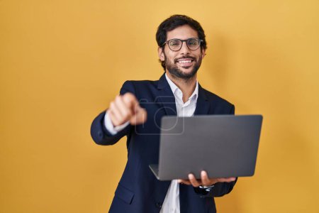 Foto de Hombre latino guapo trabajando con computadora portátil que apunta a usted y la cámara con los dedos, sonriendo positivo y alegre - Imagen libre de derechos