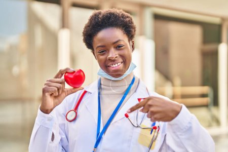 Téléchargez les photos : Femme afro-américaine portant l'uniforme de médecin tenant le cœur pointant du doigt un sourire heureux et fier - en image libre de droit