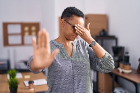 Téléchargez les photos : Femme afro-américaine travaillant au bureau portant des lunettes couvrant les yeux avec les mains et faisant un geste d'arrêt avec expression triste et peur. concept embarrassé et négatif. - en image libre de droit
