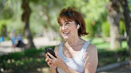 Téléchargez les photos : Femme d'âge moyen écoutant de la musique souriant au parc - en image libre de droit