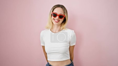Foto de Joven mujer rubia sonriendo confiada usando gafas de sol de pie sobre fondo rosa aislado - Imagen libre de derechos