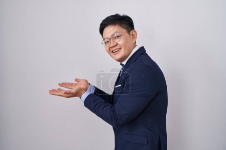 Téléchargez les photos : Jeune homme asiatique portant costume d'affaires et cravate pointant de côté avec les mains ouvertes paumes montrant l'espace de copie, présentant la publicité souriant heureux excité - en image libre de droit