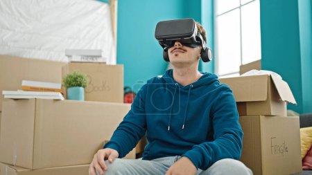 Téléchargez les photos : Jeune homme hispanique jouant à un jeu vidéo en utilisant des lunettes de réalité virtuelle à la nouvelle maison - en image libre de droit