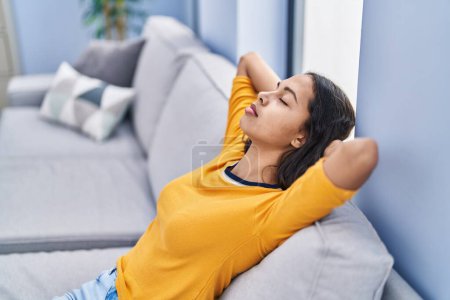 Téléchargez les photos : Jeune femme afro-américaine détendue avec les mains sur la tête assise sur le canapé à la maison - en image libre de droit