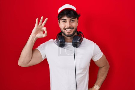 Foto de Hombre hispano con barba con sombrero de gamer y auriculares sonriendo positivo haciendo signo de ok con la mano y los dedos. expresión exitosa. - Imagen libre de derechos