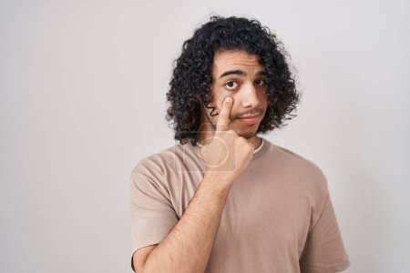 Téléchargez les photos : Homme hispanique avec les cheveux bouclés debout sur fond blanc pointant vers l'oeil vous regardant geste, expression suspecte - en image libre de droit
