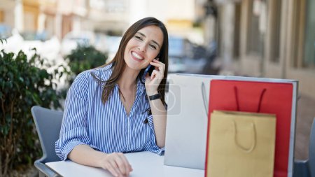 Téléchargez les photos : Jeune belle femme hispanique parlant sur smartphone assis sur la table avec des sacs à provisions à la terrasse du café - en image libre de droit