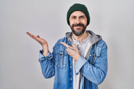 Téléchargez les photos : Jeune homme hispanique avec des tatouages portant un bonnet de laine étonné et souriant à la caméra tout en présentant avec la main et pointant du doigt. - en image libre de droit