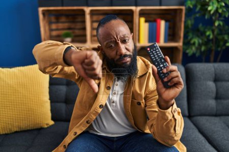 Foto de Hombre afroamericano sosteniendo control remoto de televisión con la cara enojada, signo negativo que muestra aversión con los pulgares hacia abajo, concepto de rechazo - Imagen libre de derechos