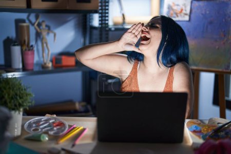 Téléchargez les photos : Jeune fille moderne avec les cheveux bleus assis au studio d'art avec ordinateur portable la nuit criant et criant fort à côté avec la main sur la bouche. concept de communication. - en image libre de droit