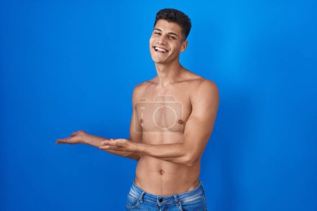 Téléchargez les photos : Jeune homme hispanique debout torse nu sur fond bleu invitant à entrer souriant naturel avec la main ouverte - en image libre de droit