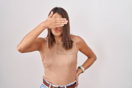 Téléchargez les photos : Jeune femme hispanique debout sur fond blanc couvrant les yeux avec la main, l'air grave et triste. concept de non-visibilité, de dissimulation et de rejet - en image libre de droit