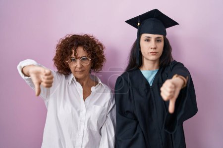 Photo for Hispanic mother and daughter wearing graduation cap and ceremony robe looking unhappy and angry showing rejection and negative with thumbs down gesture. bad expression. - Royalty Free Image