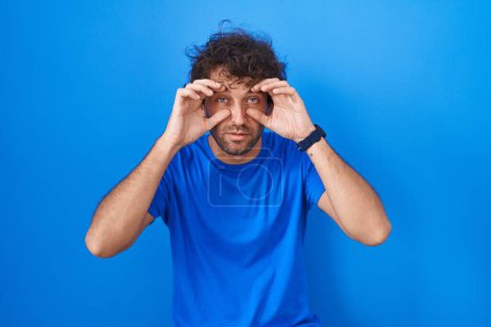 Foto de Hispanic young man standing over blue background trying to open eyes with fingers, sleepy and tired for morning fatigue - Imagen libre de derechos