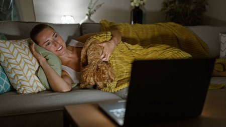 Téléchargez les photos : Jeune femme caucasienne avec chien regarder film sur ordinateur portable couché sur canapé souriant à la maison - en image libre de droit