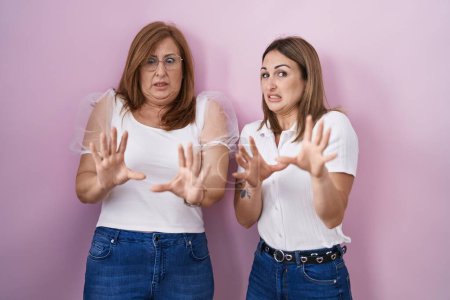 Téléchargez les photos : Mère et fille hispaniques portant un t-shirt blanc décontracté sur fond rose effrayés et terrifiés par l'expression de la peur arrêtent geste avec les mains, criant en état de choc. concept de panique. - en image libre de droit