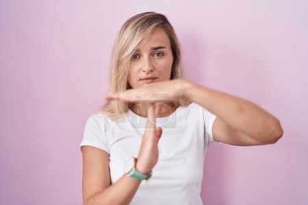 Photo for Young blonde woman standing over pink background doing time out gesture with hands, frustrated and serious face - Royalty Free Image