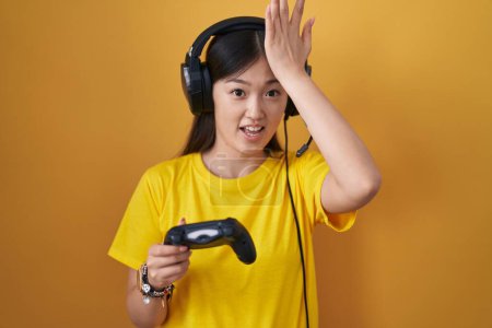 Foto de Mujer joven china jugando el controlador de retención de videojuegos sorprendido con la mano en la cabeza por error, recuerde el error. olvidado, mal concepto de memoria. - Imagen libre de derechos