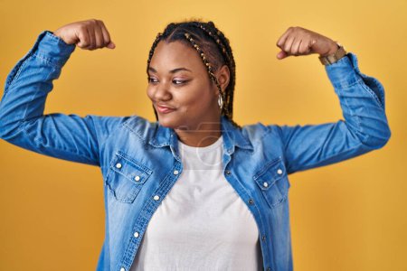 Téléchargez les photos : Femme afro-américaine avec des tresses debout sur fond jaune montrant les muscles des bras souriant fier. concept de fitness. - en image libre de droit