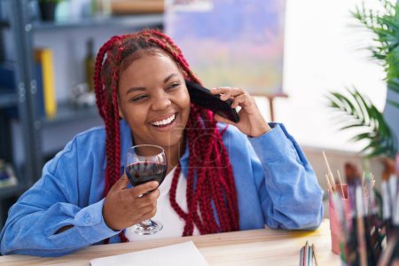 Téléchargez les photos : Artiste afro-américaine parlant sur smartphone boire du vin au studio d'art - en image libre de droit