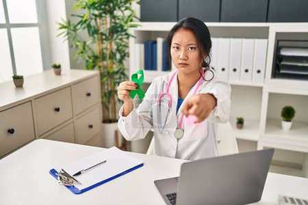 Photo for Young asian doctor woman holding support green ribbon pointing with finger to the camera and to you, confident gesture looking serious - Royalty Free Image