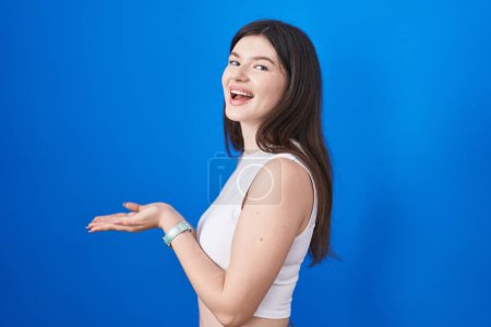 Téléchargez les photos : Jeune femme caucasienne debout sur fond bleu pointant de côté avec les mains ouvertes paumes montrant l'espace de copie, présentant la publicité souriant heureux excité - en image libre de droit