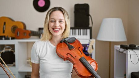 Foto de Joven mujer rubia músico sosteniendo violín sonriendo en el estudio de música - Imagen libre de derechos