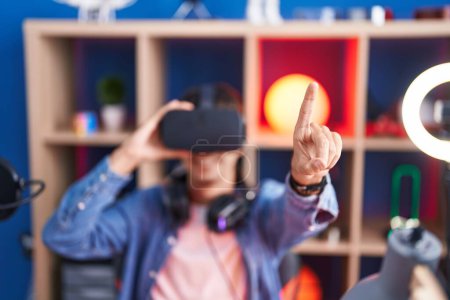 Téléchargez les photos : Jeune homme hispanique streamer jouer à un jeu vidéo en utilisant des lunettes de réalité virtuelle à la salle de jeu - en image libre de droit