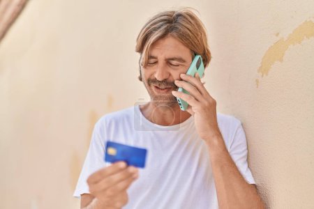Foto de Joven hombre rubio hablando en el teléfono inteligente con tarjeta de crédito en la calle - Imagen libre de derechos
