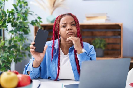 Foto de Mujer afroamericana con cabello trenzado sosteniendo teléfono inteligente que muestra la cara seria de la pantalla en blanco pensando en la pregunta con la mano en la barbilla, reflexivo sobre la idea confusa - Imagen libre de derechos