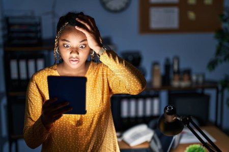Foto de Mujer afroamericana con trenzas trabajando en la oficina por la noche con la tableta loca y asustada con las manos en la cabeza, asustada y sorprendida de shock con la boca abierta - Imagen libre de derechos