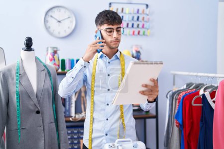 Téléchargez les photos : Jeune homme hispanique tailleur parler sur smartphone regarder carnet à tailleur boutique - en image libre de droit