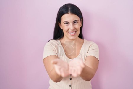 Téléchargez les photos : Jeune femme hispanique debout sur fond rose souriant avec les mains paumes ensemble recevant ou donnant geste. maintien et protection - en image libre de droit