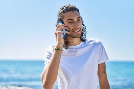 Téléchargez les photos : Jeune homme hispanique souriant confiant parler sur le smartphone au bord de la mer - en image libre de droit