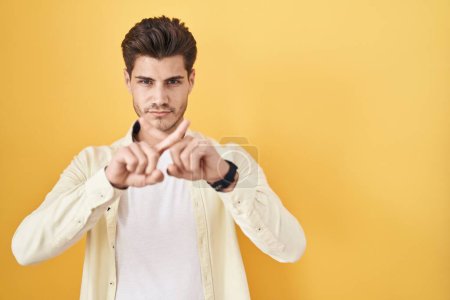 Foto de Joven hombre hispano de pie sobre fondo amarillo expresión rechazo cruzando dedos haciendo signo negativo - Imagen libre de derechos