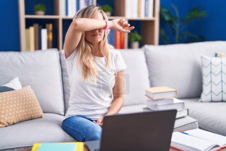 Téléchargez les photos : Jeune femme blonde étudiant à l'aide d'un ordinateur portable à la maison couvrant les yeux avec le bras, l'air grave et triste. concept de non-visibilité, de dissimulation et de rejet - en image libre de droit
