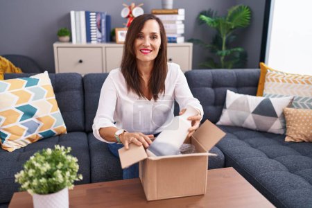 Photo for Young beautiful hispanic woman unpacking plant jar of cardboard box sitting on table at home - Royalty Free Image